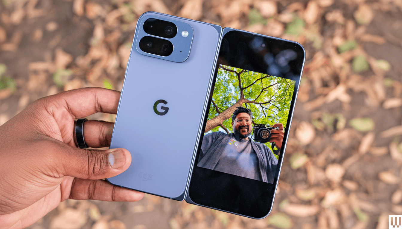 A hand holding a light blue foldable smartphone, displaying a selfie of a man holding a camera on its screen, against a blurred background of autumn leaves.