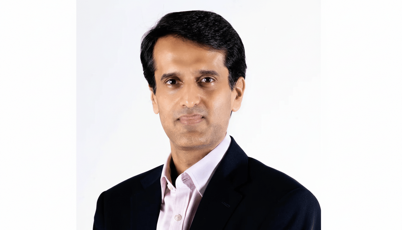 A professional headshot of a man in a dark suit and pink shirt, resized to a 16:9 aspect ratio with the original white background maintained.