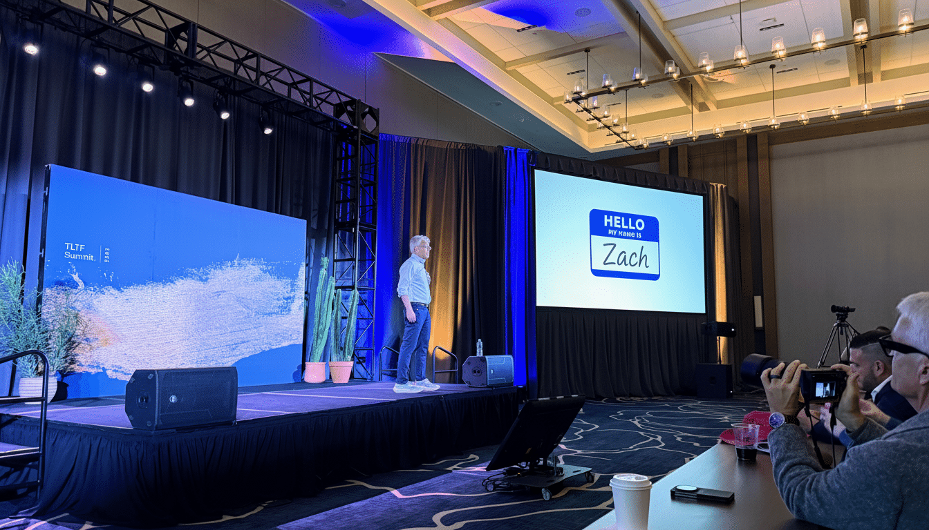 A man stands on a stage presenting, with a large screen to his right displaying a Hello, my name is Zach nametag.