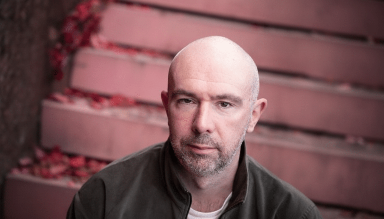 A bald man with a beard looking directly at the camera, with a blurred background of pink stairs adorned with flower petals.