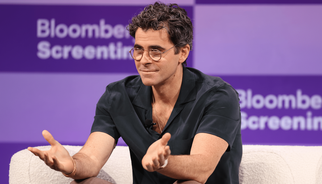 A man with curly dark hair and glasses, wearing a dark short-sleeved shirt, sits on a white textured couch with his hands open in a gesturing manner. The background is purple with Bloomberg Screentime text visible.