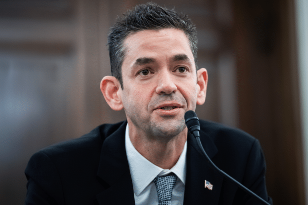 A man in a suit and tie speaking into a microphone, resized to a 16:9 aspect ratio.