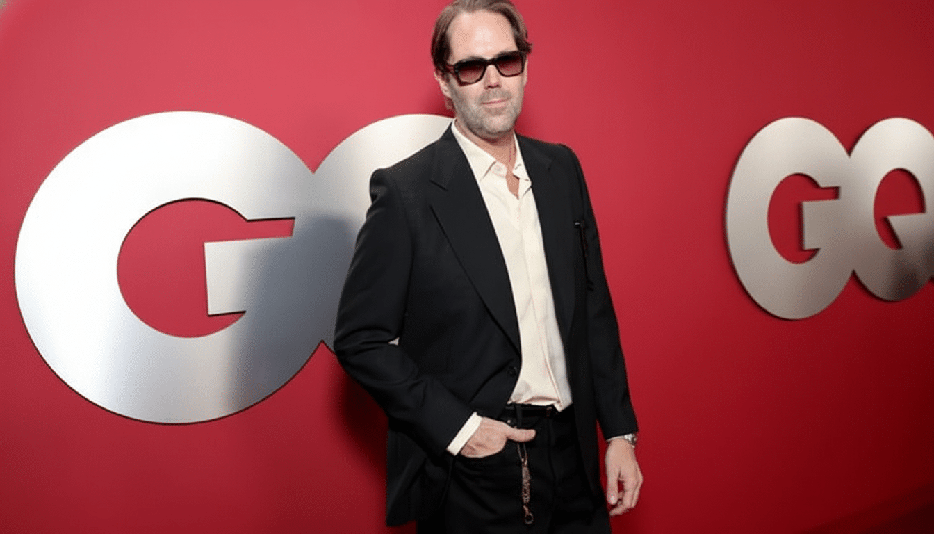 A man in a black suit and sunglasses stands in front of a red wall with a large silver GQ logo.
