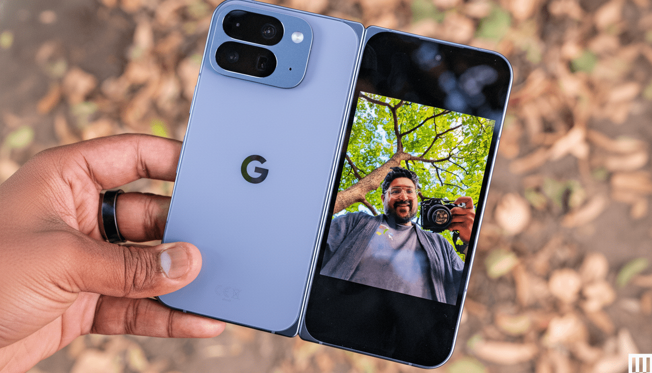 A hand holding a light blue Google Pixel Fold phone, with the screen displaying a selfie of a man holding a camera, against a blurred background of autumn leaves.