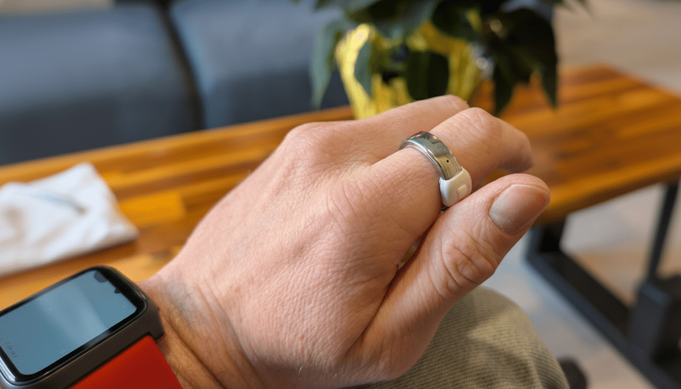 A hand wearing a silver smart ring with a white sensor, with a smartwatch on the wrist, against a blurred background of a wooden table and a plant.