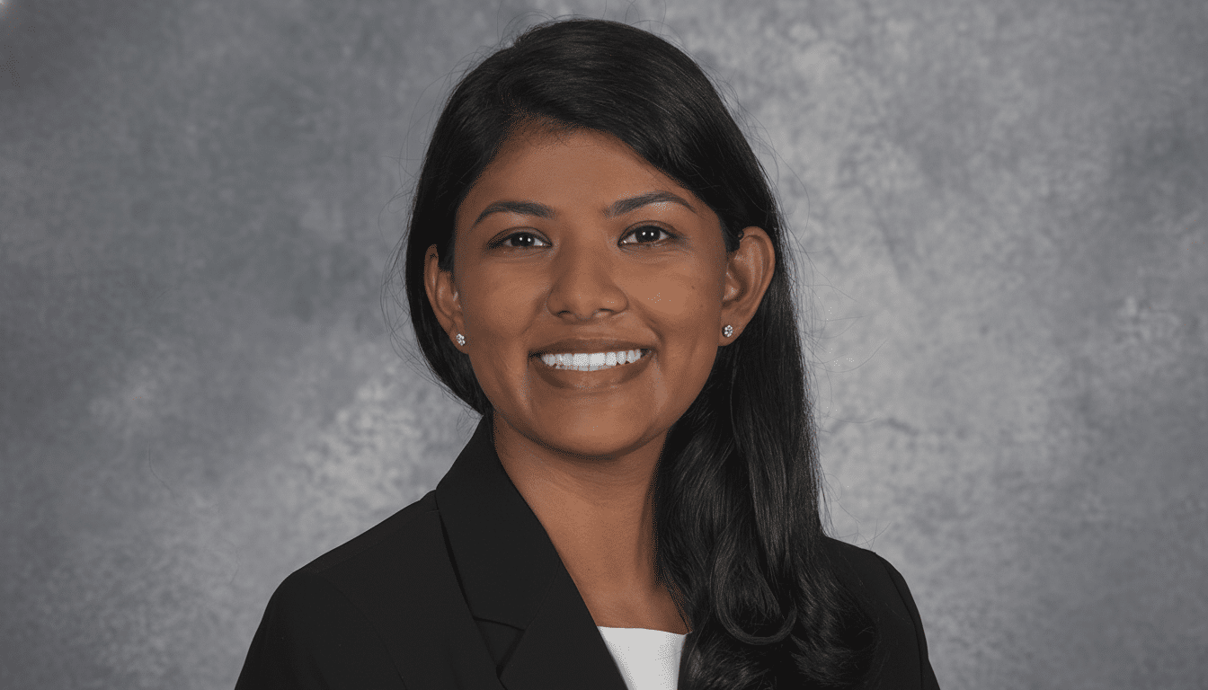 A professional headshot of a woman with dark hair and a black blazer, smiling against a grey background.