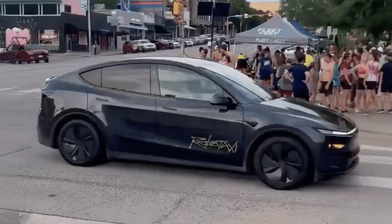 A dark grey Tesla with ROBOTAXI written on its side, driving on a street with people gathered in the background.