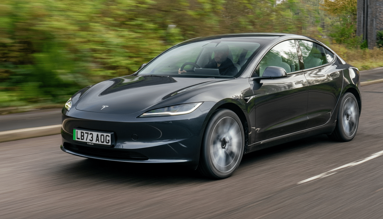 A dark grey Tesla Model 3 driving on a road with blurred green foliage in the background.