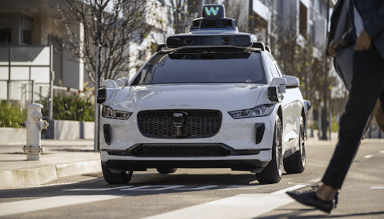 A white self-driving car with a Waymo logo on top, stopped at a crosswalk with a person walking in the foreground.