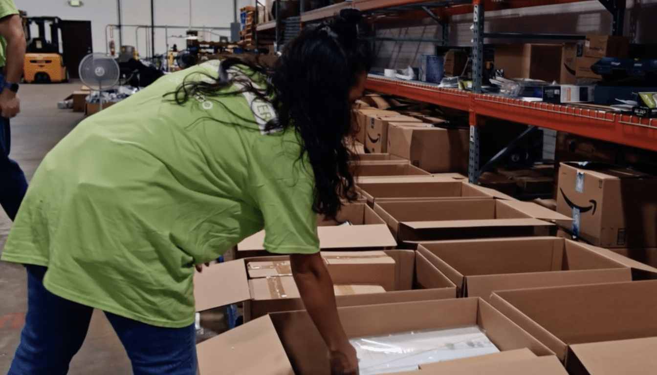 A person in a green shirt and blue jeans is placing an item into a cardboard box in a warehouse setting.