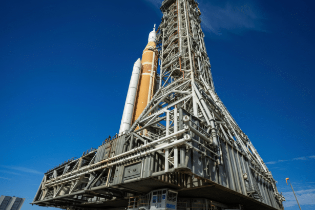 A large rocket, the Space Launch System, stands on a mobile launcher against a clear blue sky.