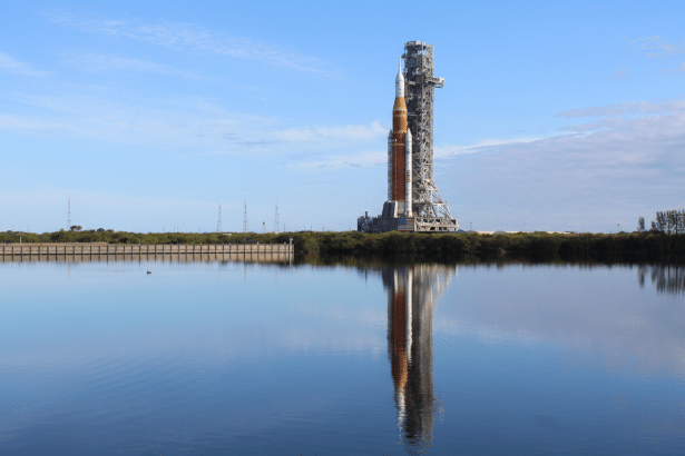 A large rocket on a launchpad, reflected in the calm water in the foreground.