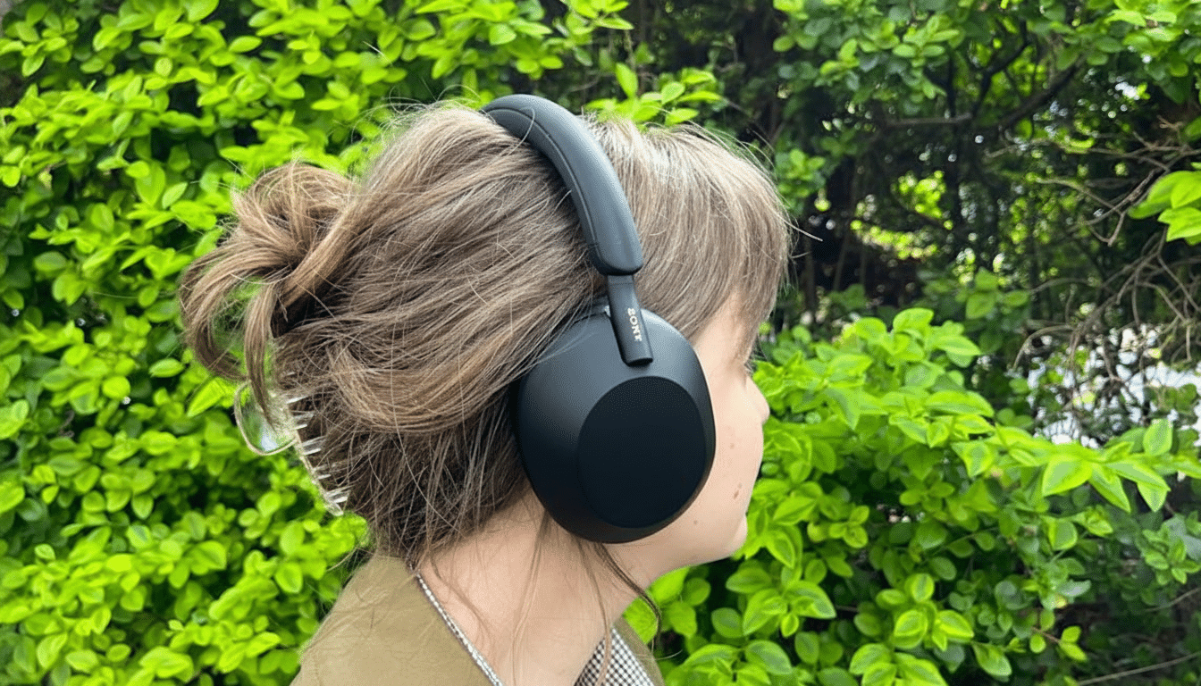A person with brown hair in a bun, wearing black Sony over-ear headphones, stands in front of a vibrant green leafy background.