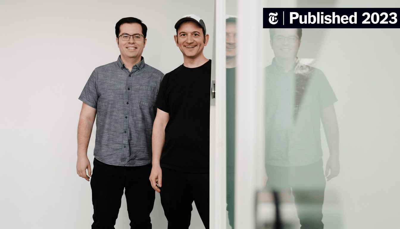 Two men smiling at the camera, one wearing glasses and a grey shirt, the other wearing a black shirt and a cap, with a reflection of another person in the background.