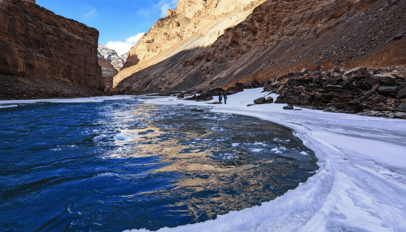 A wide shot of a river flowing through a canyon, with snow and ice on the banks and two figures standing in the distance.