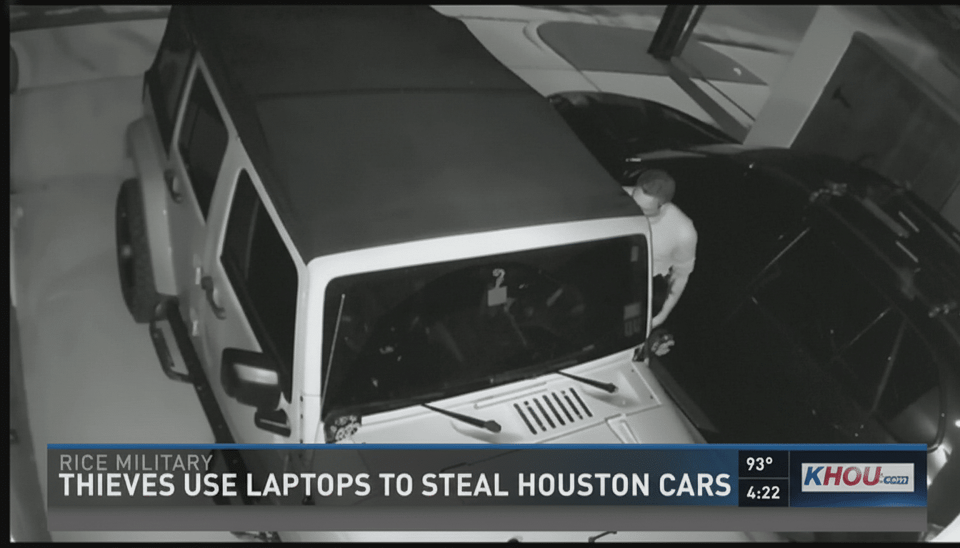 A black and white security camera still shows a person in a light-colored shirt leaning into the engine bay of a dark car, parked next to a white Jeep. A news banner at the bottom reads THIEVES USE LAPTOPS TO STEAL HOUSTON CARS.