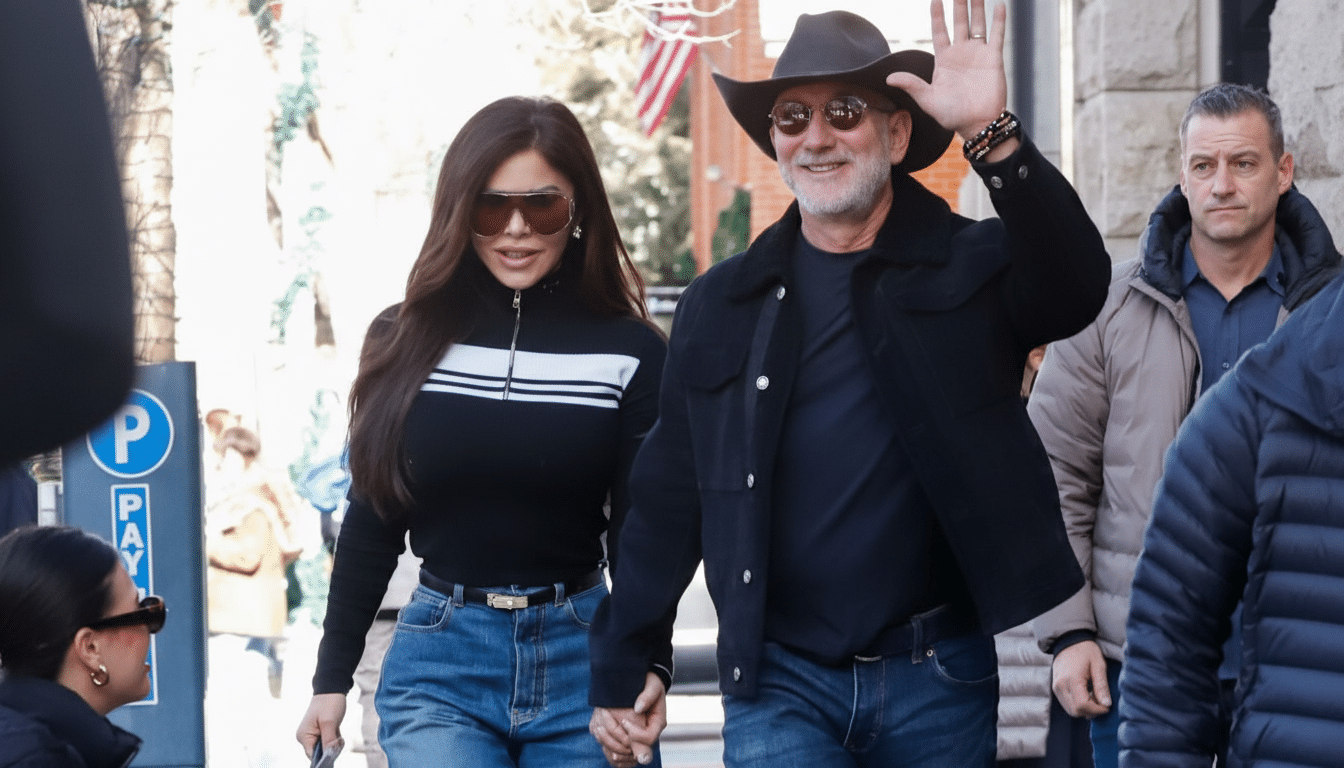 A man in a cowboy hat and sunglasses waves while holding hands with a woman in sunglasses and a black and white striped top, walking down a street.