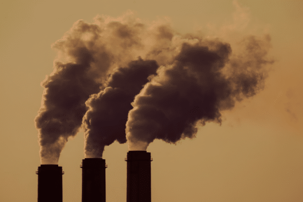 Three industrial smokestacks emit plumes of dark smoke against a warm, hazy sky, resized to a 16:9 aspect ratio.