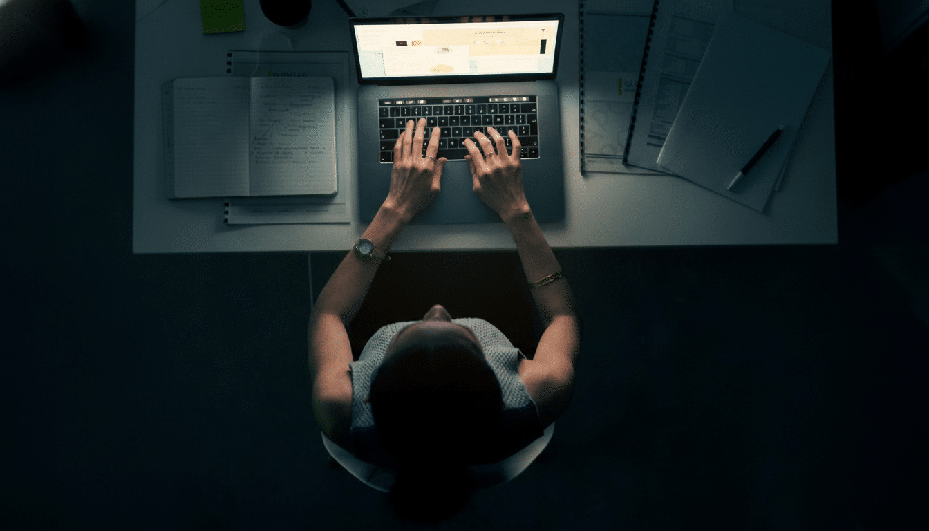 An overhead shot of a person working on a laptop at a desk, with notebooks and papers scattered around.