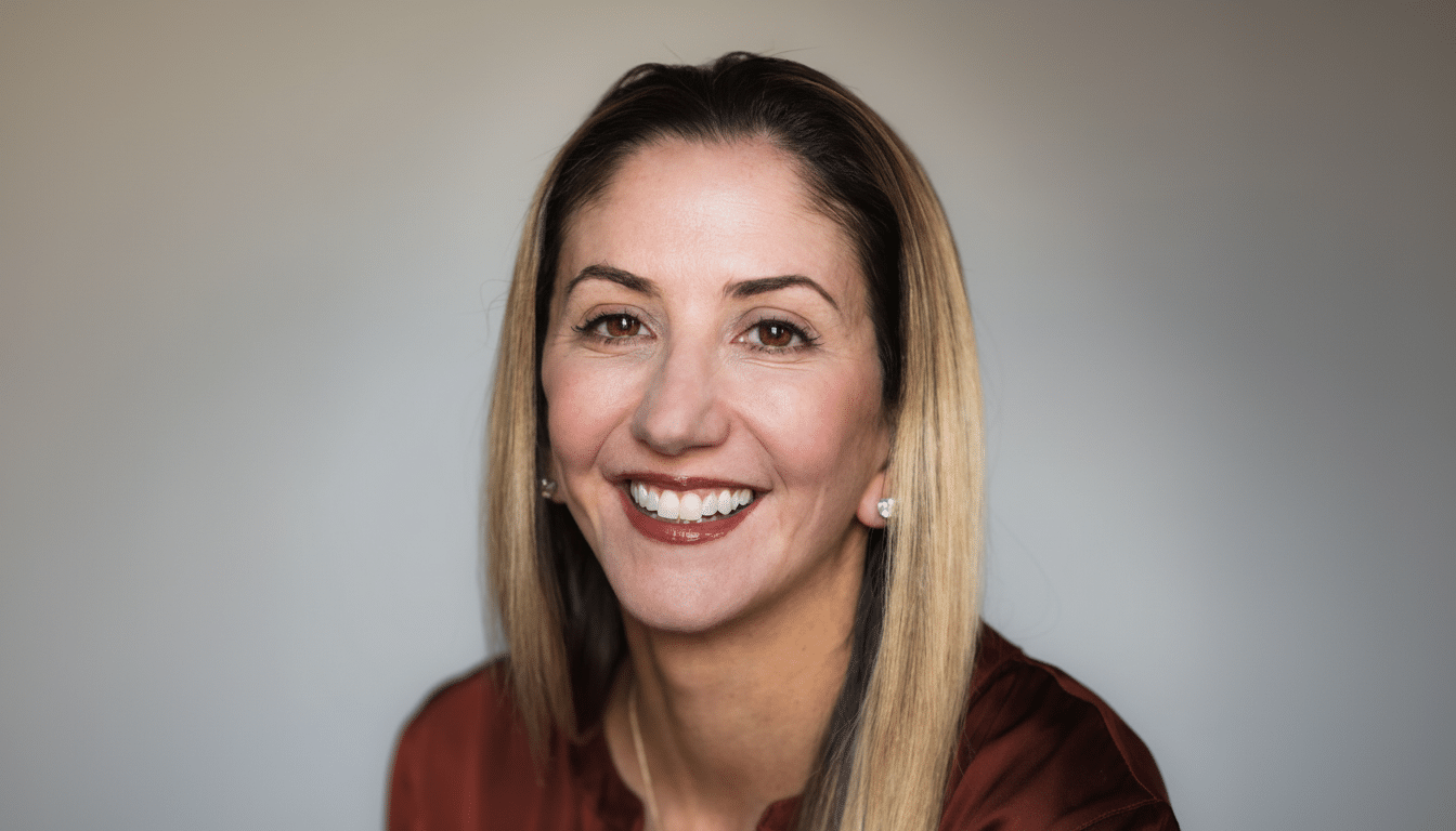 A professional headshot of a smiling woman with long blonde hair, wearing a rust-colored top, against a light gray background.