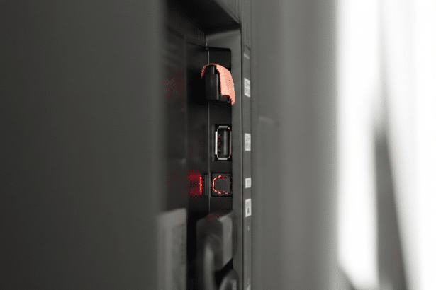 A close-up, side view of the back of a dark gray television, showing various ports including a USB port, an optical audio port with a red light, and another port with an orange cover. The background is a soft, out-of-focus white and gray.