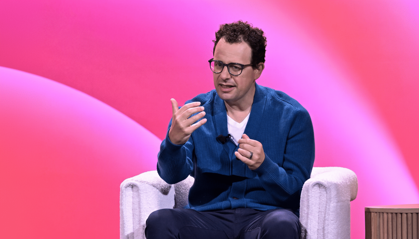 A man with dark curly hair and glasses, wearing a blue cardigan over a white t-shirt and dark pants, sits in a white textured armchair against a vibrant pink and red gradient background. He is gesturing with both hands as if speaking.