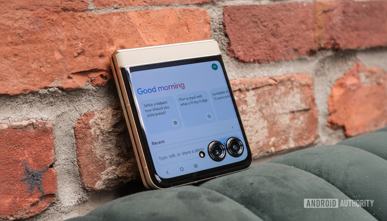 A gold and white foldable smartphone displaying Good morning on its screen, resting against a brick wall and a dark green cushioned surface.