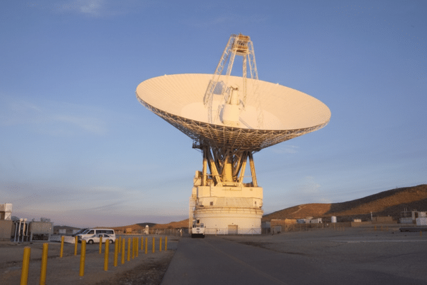 A large white radio telescope dish stands against a clear blue sky with a desert landscape in the background.