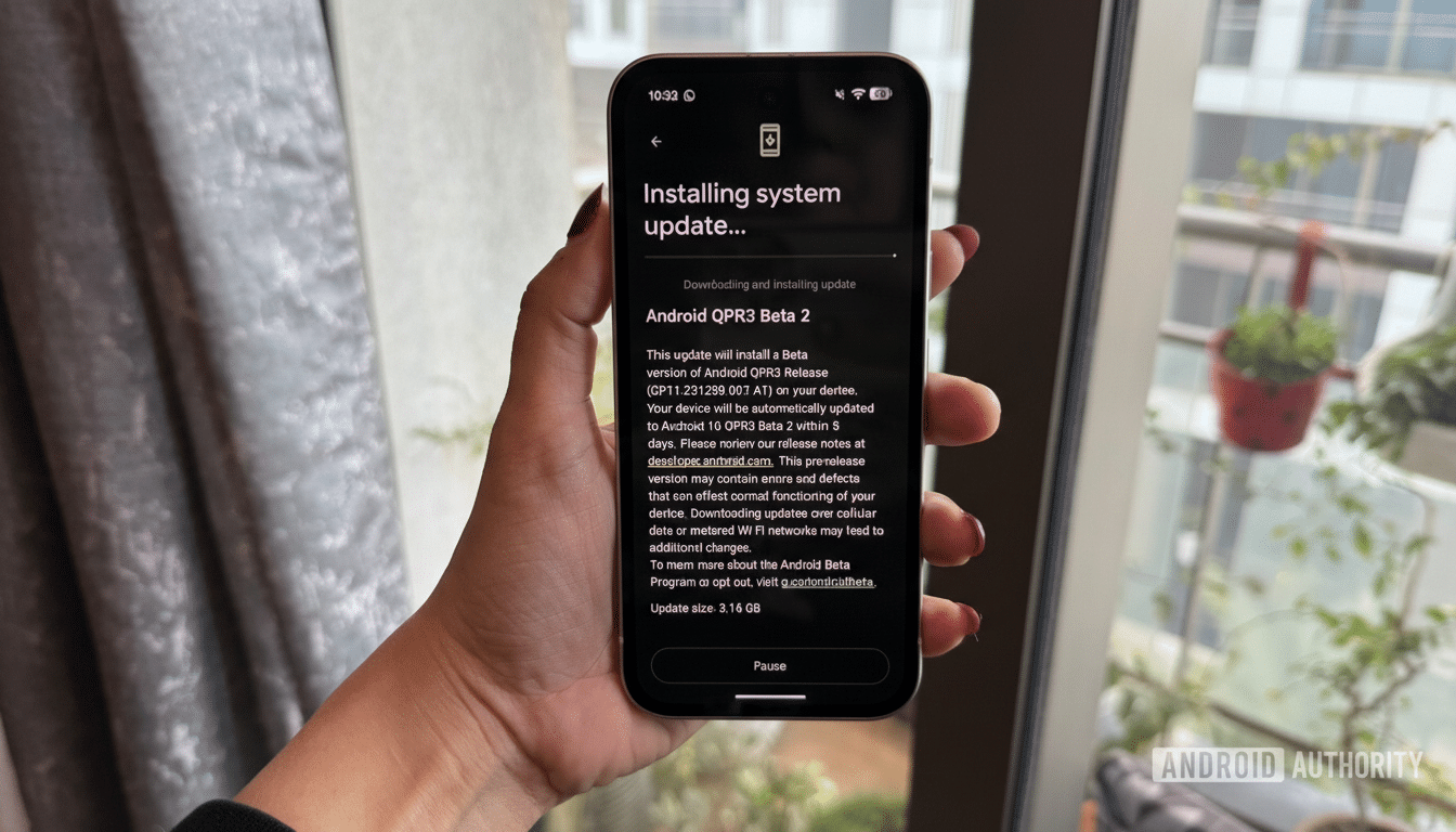 A hand holding a smartphone displaying an Installing system update... screen for Android QPR3 Beta 2.