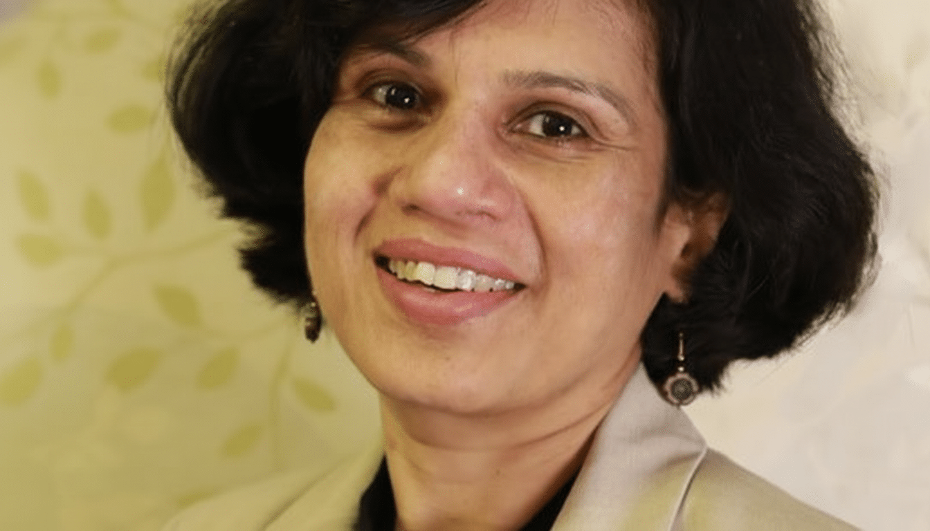 A professional headshot of a smiling woman with short dark hair, wearing a light-colored blazer, against a subtly patterned background.