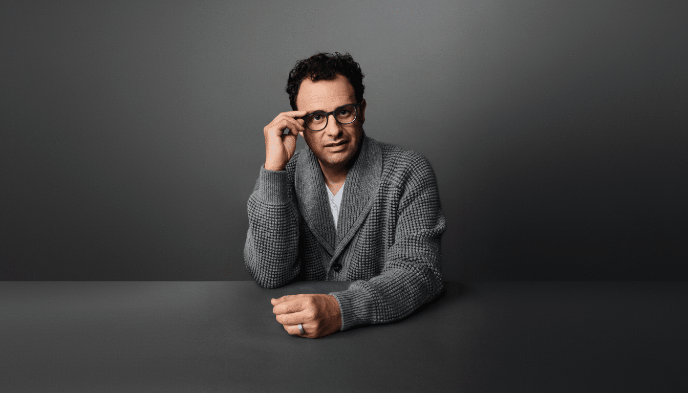 A man with dark curly hair and glasses, wearing a grey cardigan and white t-shirt, sits at a dark table against a grey background, looking directly at the viewer.