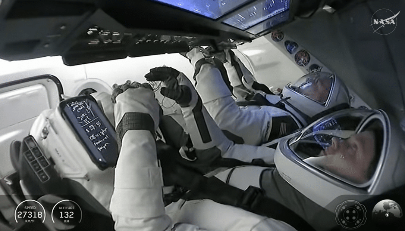 Two astronauts in white spacesuits are inside a spacecraft, with one looking at a tablet displaying data.