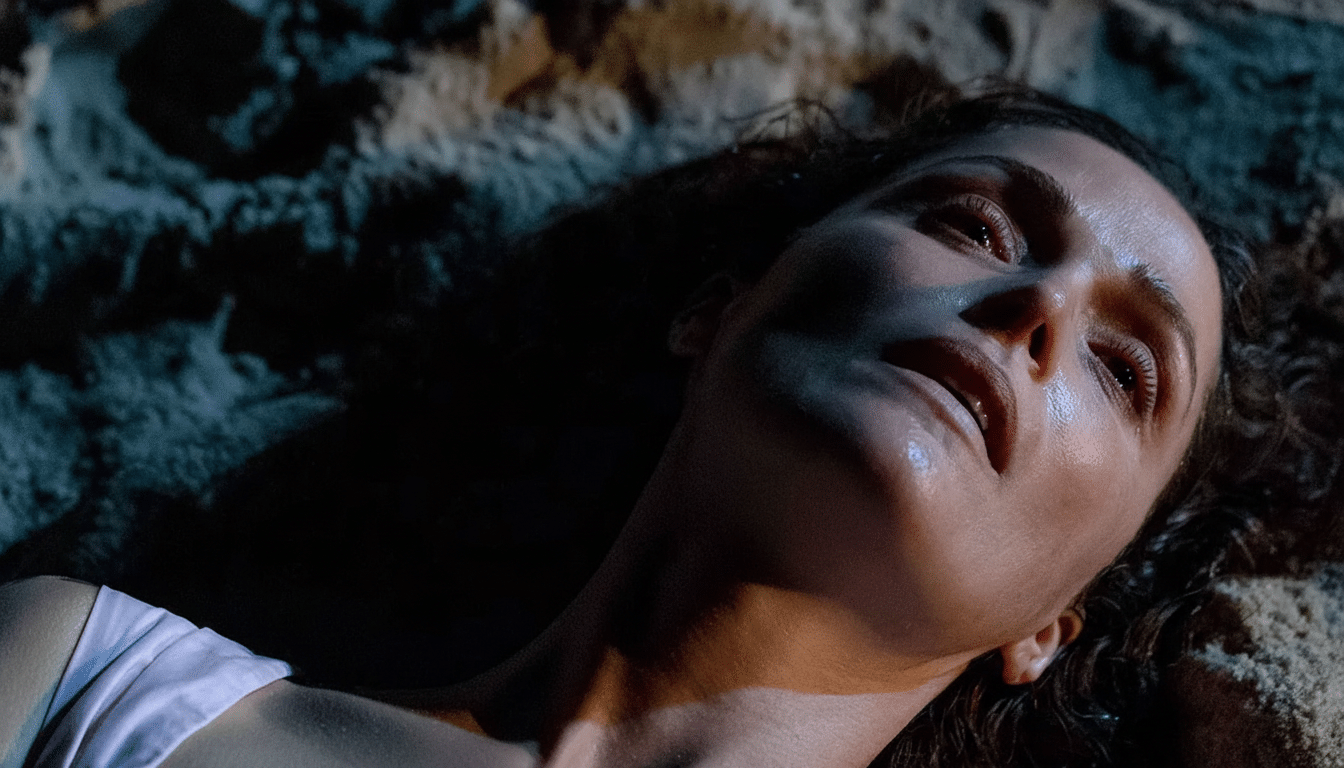 A woman with dark hair and a light pink top lies on sand, looking upwards with a pensive expression.