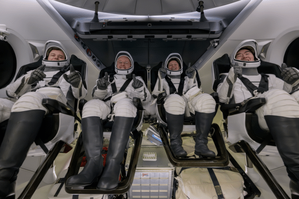 Four astronauts in white spacesuits are seated in a spacecraft, giving thumbs up.