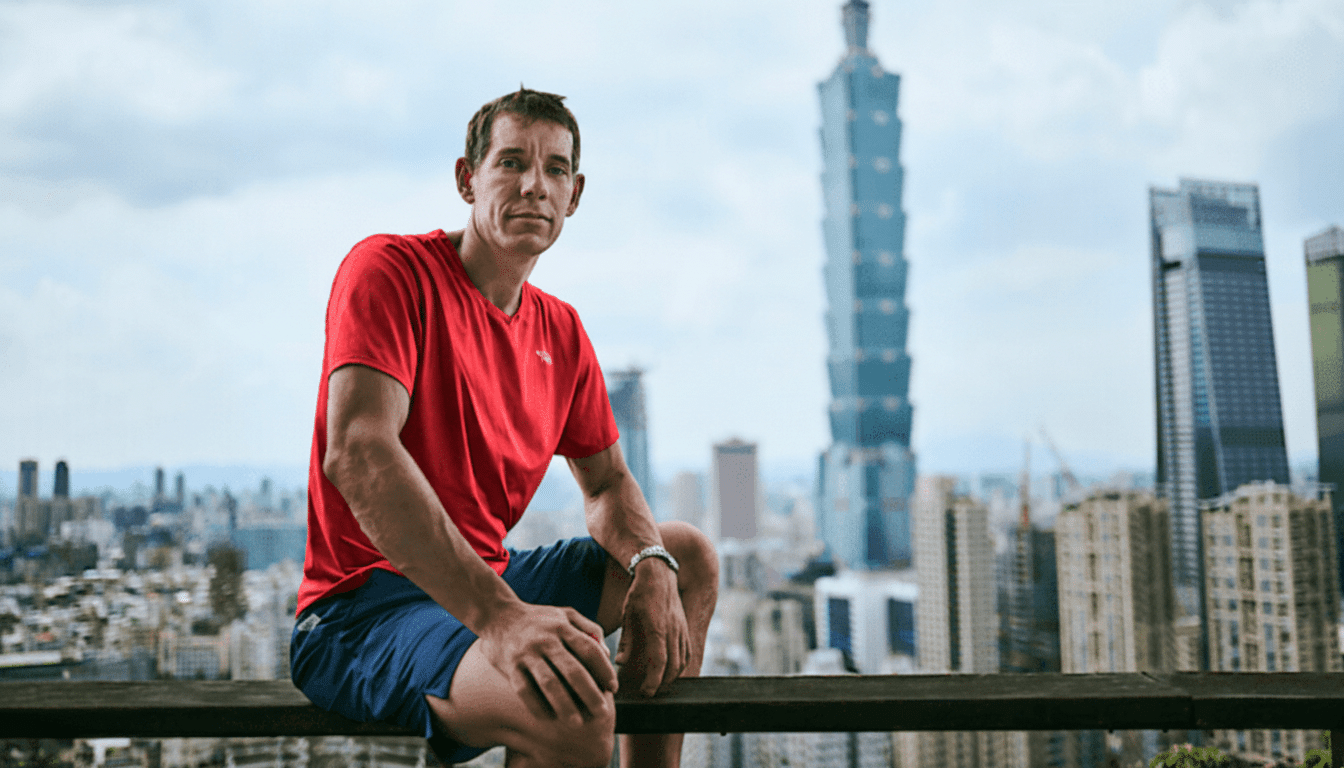 A man in a red shirt and blue shorts sits on a ledge with a city skyline and a tall skyscraper in the background.