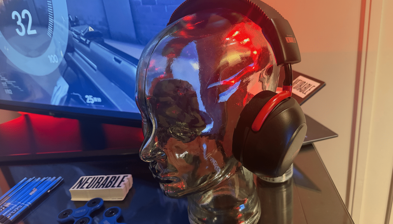 A pair of black and red headphones on a clear glass head display, with a computer monitor showing a game in the background.