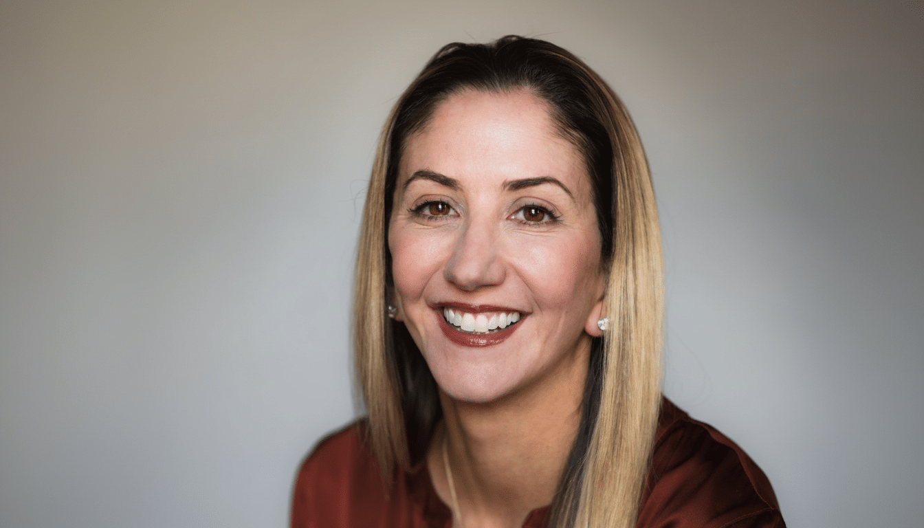 A professional headshot of a smiling woman with long blonde hair, wearing a rust-colored top, against a light gray background.