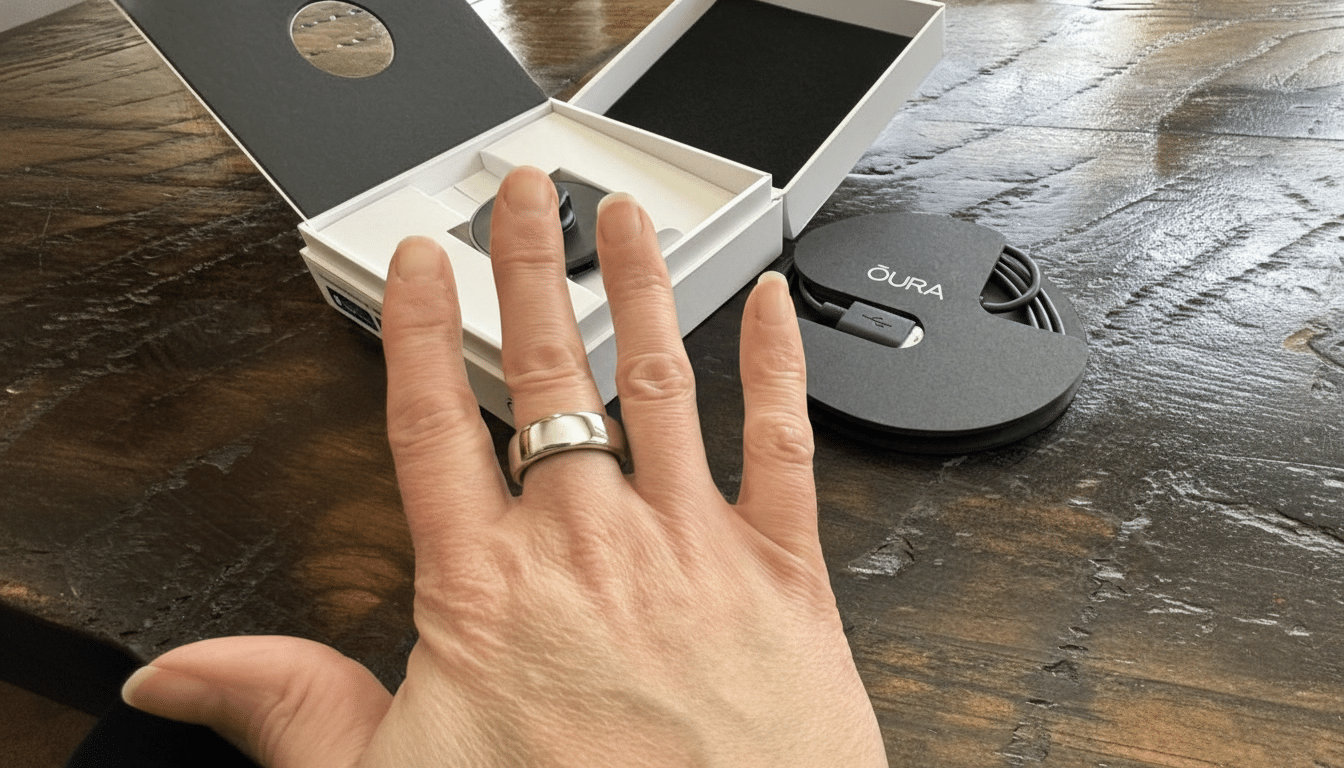 A hand wearing a silver Oura Ring, with the Oura Ring box and charging cable visible in the background on a wooden table.