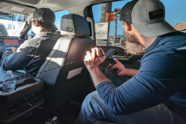 Two men in a vehicle, one looking out the window and the other working on a laptop.