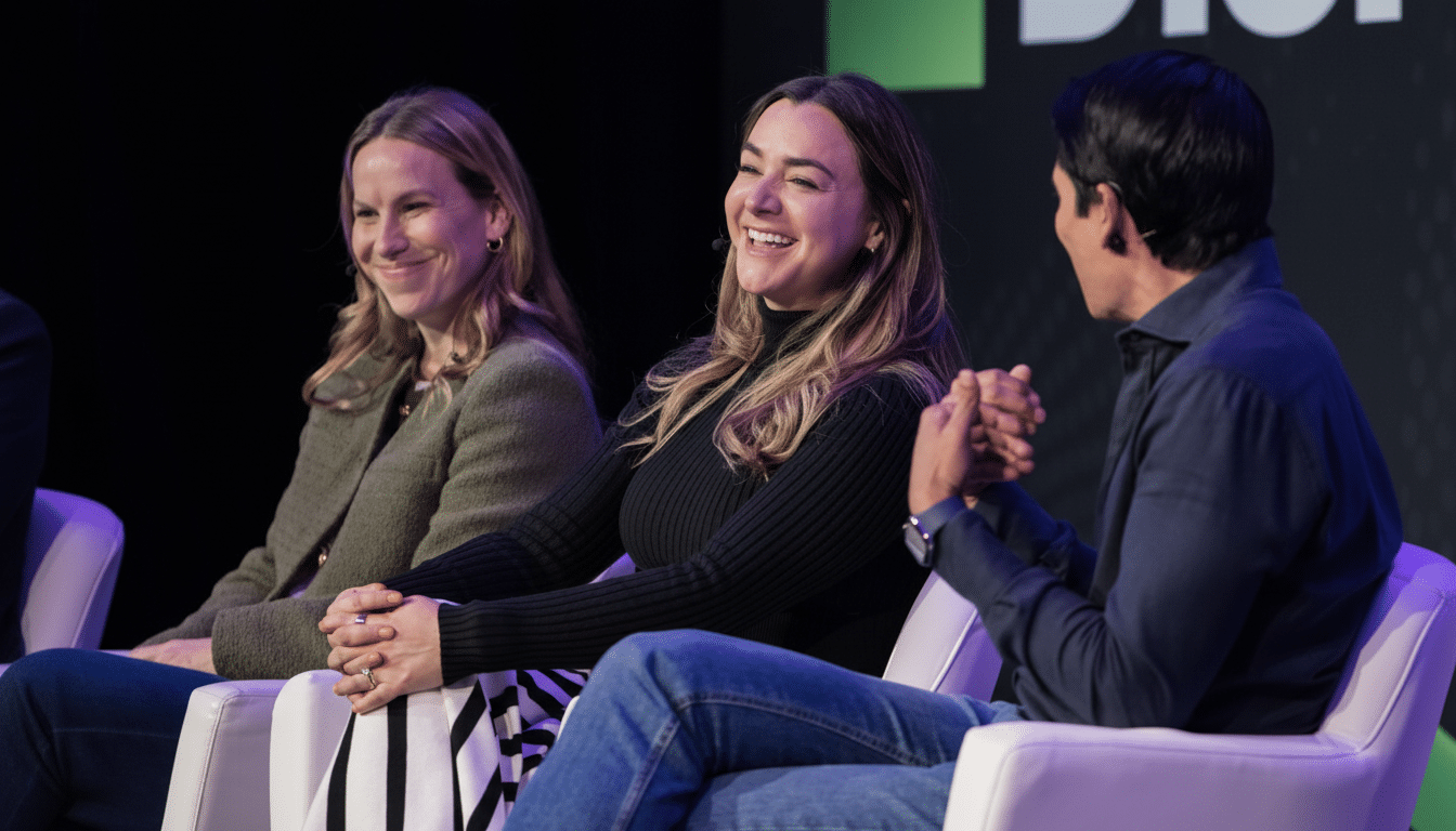 Three people, two women and one man, are seated on a stage, engaged in conversation and smiling. The woman in the center is laughing, while the woman to her left is smiling subtly. The man on the right is looking towards the center woman, with his hands clasped. The background features a dark screen with a green and white logo.