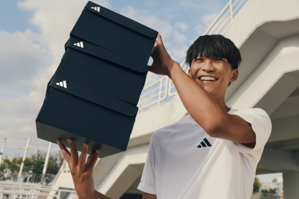 A smiling man in a white Adidas shirt holding a stack of three black Adidas shoeboxes outdoors.
