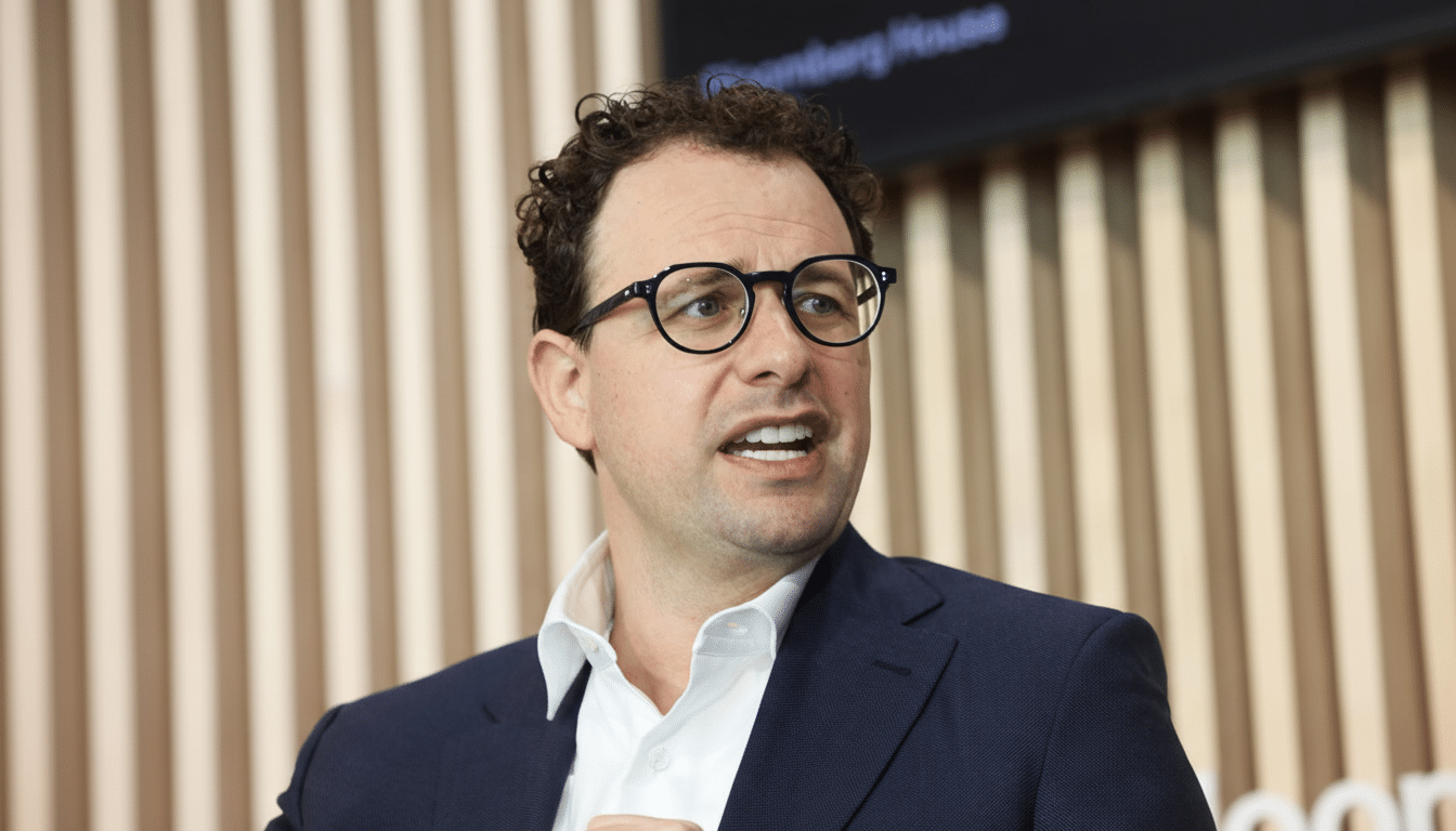 A man with curly brown hair and glasses, wearing a dark suit and white shirt, speaking at an event with a wooden slatted wall in the background.