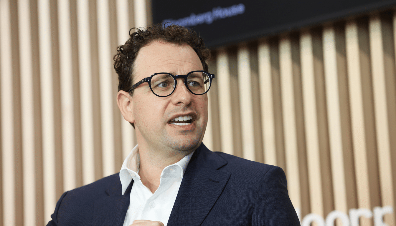 A man with curly brown hair and glasses, wearing a dark suit and white shirt, speaking with an open mouth.