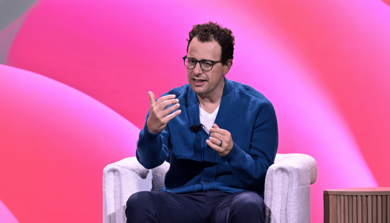 A man with curly hair and glasses, wearing a blue cardigan and white t-shirt, sits in a white chair against a pink background, gesturing with his hands.