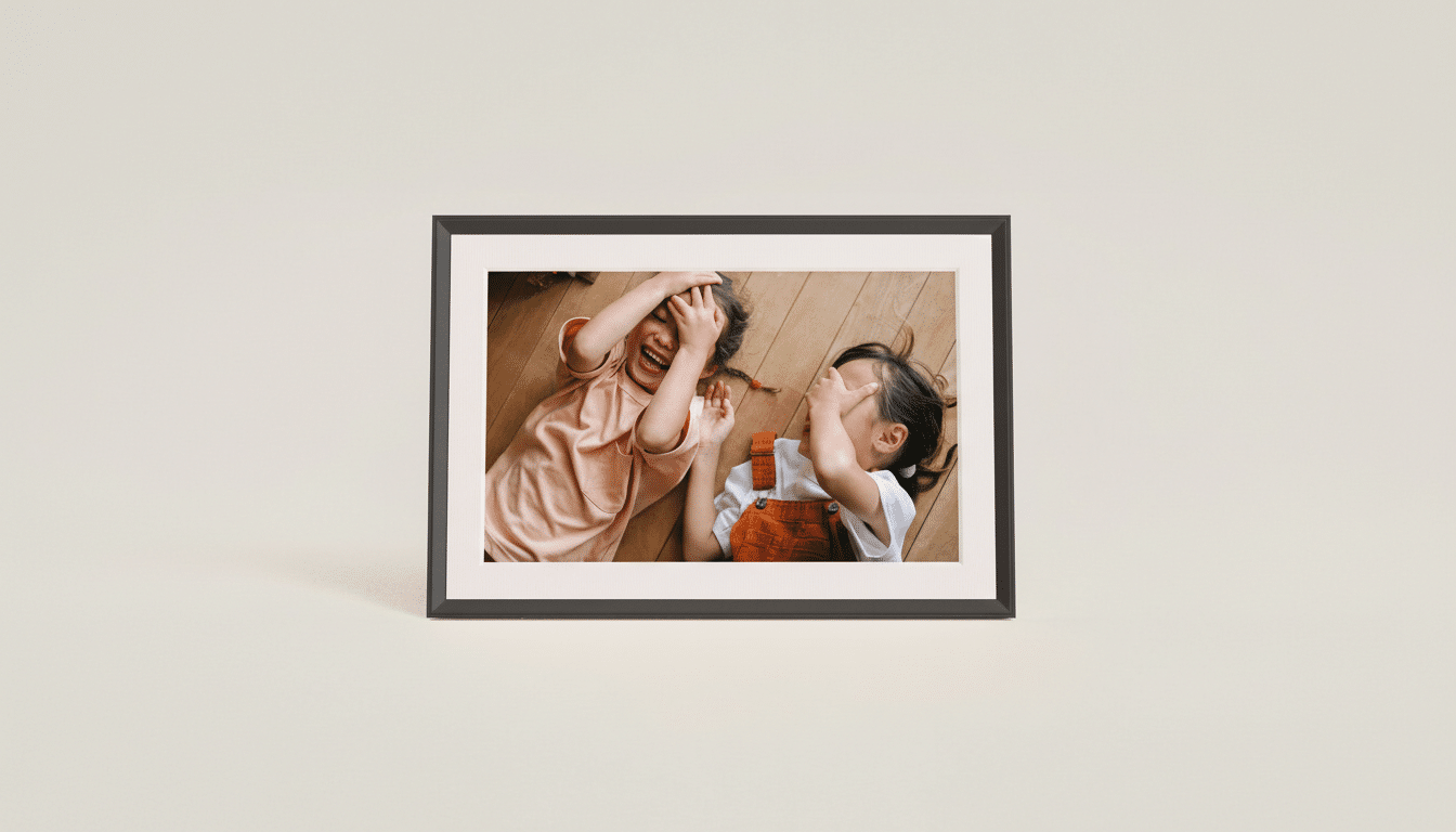 A framed photo of two young children lying on a wooden floor, laughing and covering their faces, presented on a light beige background.