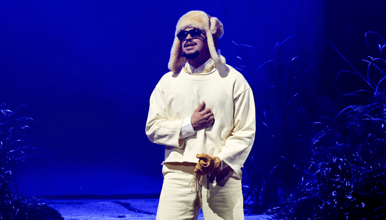 A man wearing a cream-colored outfit, a fur hat, and sunglasses stands on a stage with a blue background.