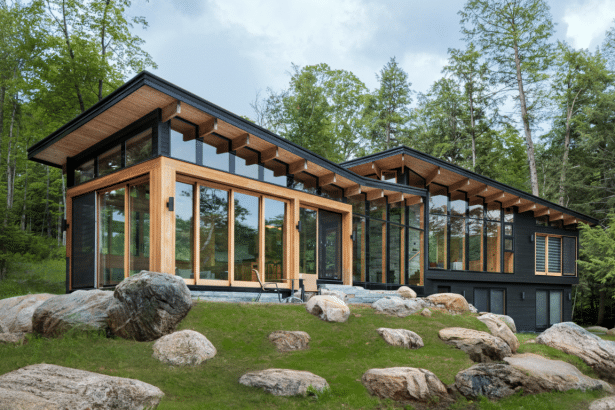 A modern house with large windows and a wooden frame, surrounded by trees and rocks, resized to a 16:9 aspect ratio.