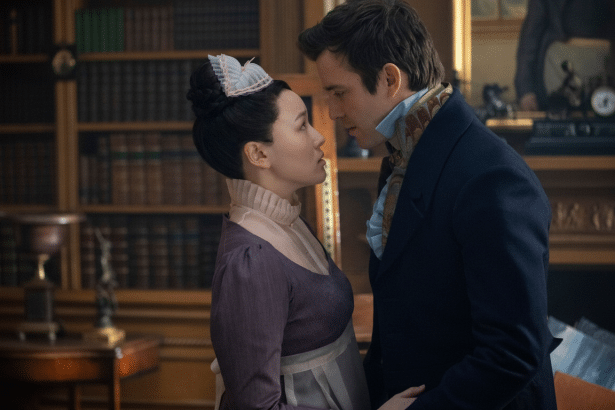 A man and a woman in period clothing gaze at each other intently in a room with bookshelves.