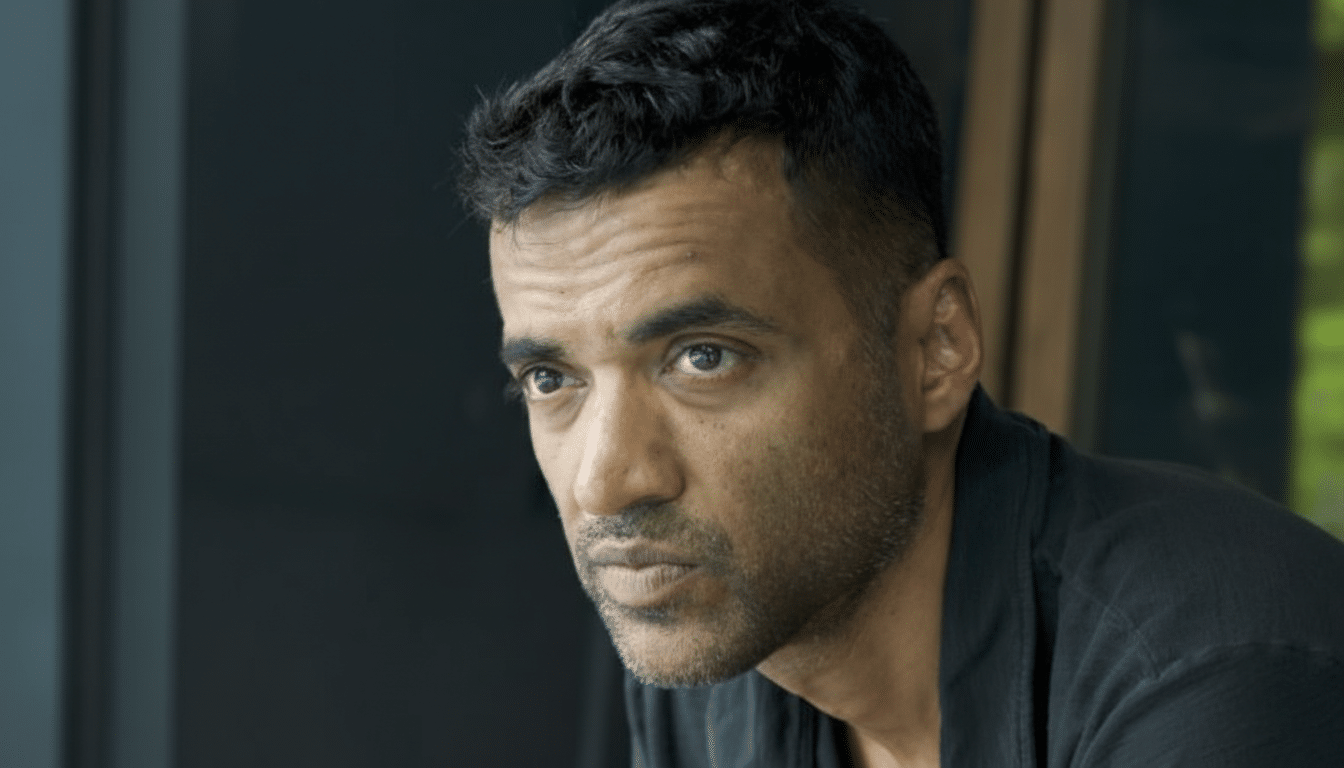 A man with short dark hair and a beard, wearing a dark shirt, looking slightly to the left with a serious expression.