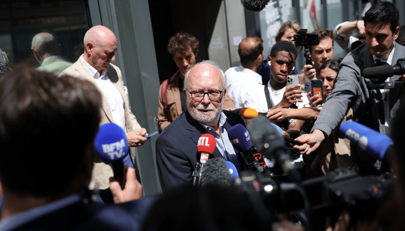 A man with glasses and a beard speaks into several microphones held by reporters, surrounded by a crowd of people.